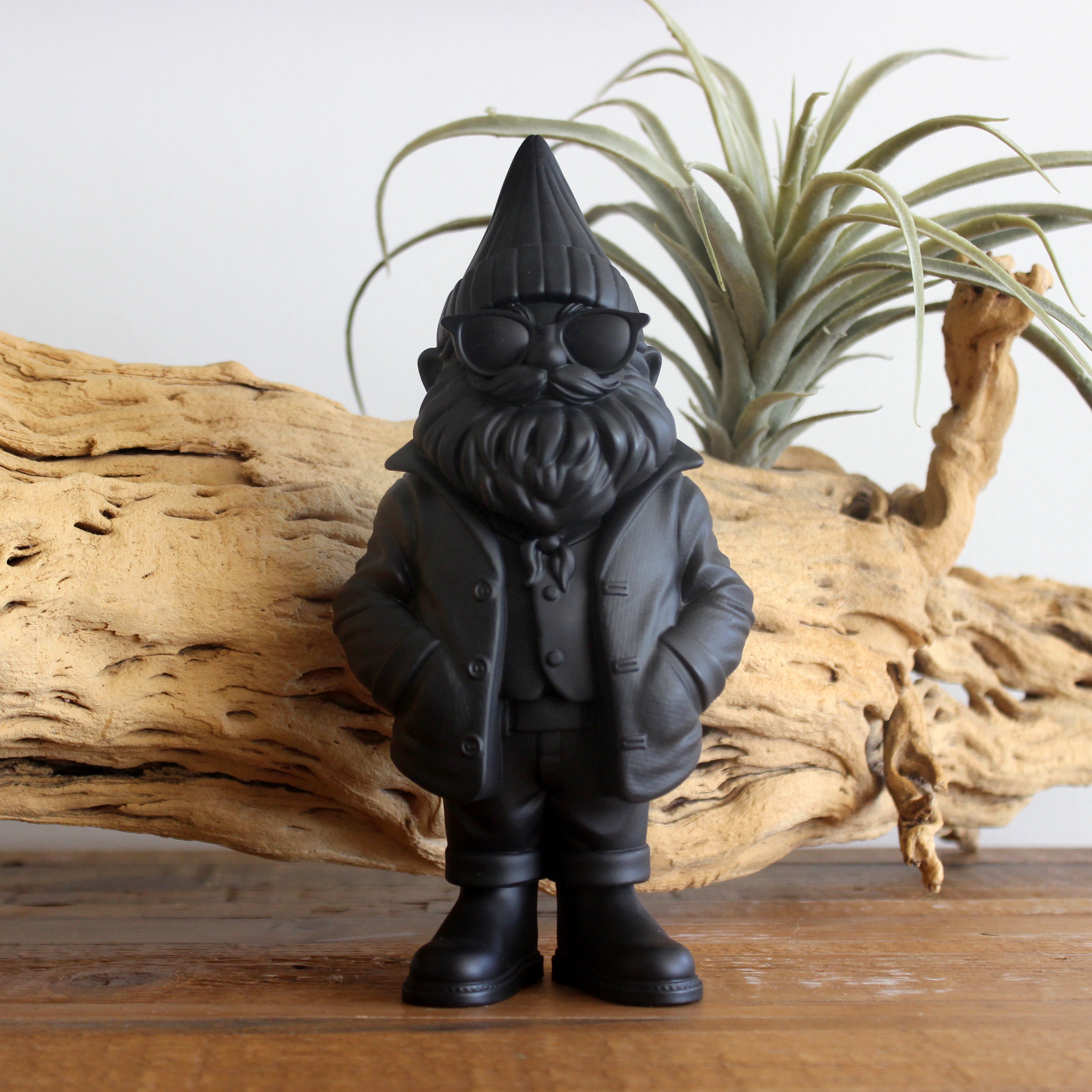Black gnome figurine with a hat and glasses on a wooden surface with driftwood and a plant in the background.